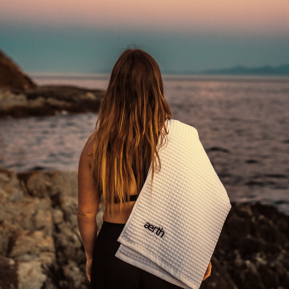 Person standing on a rocky beach with a towel draped over their shoulder, looking out at the ocean.