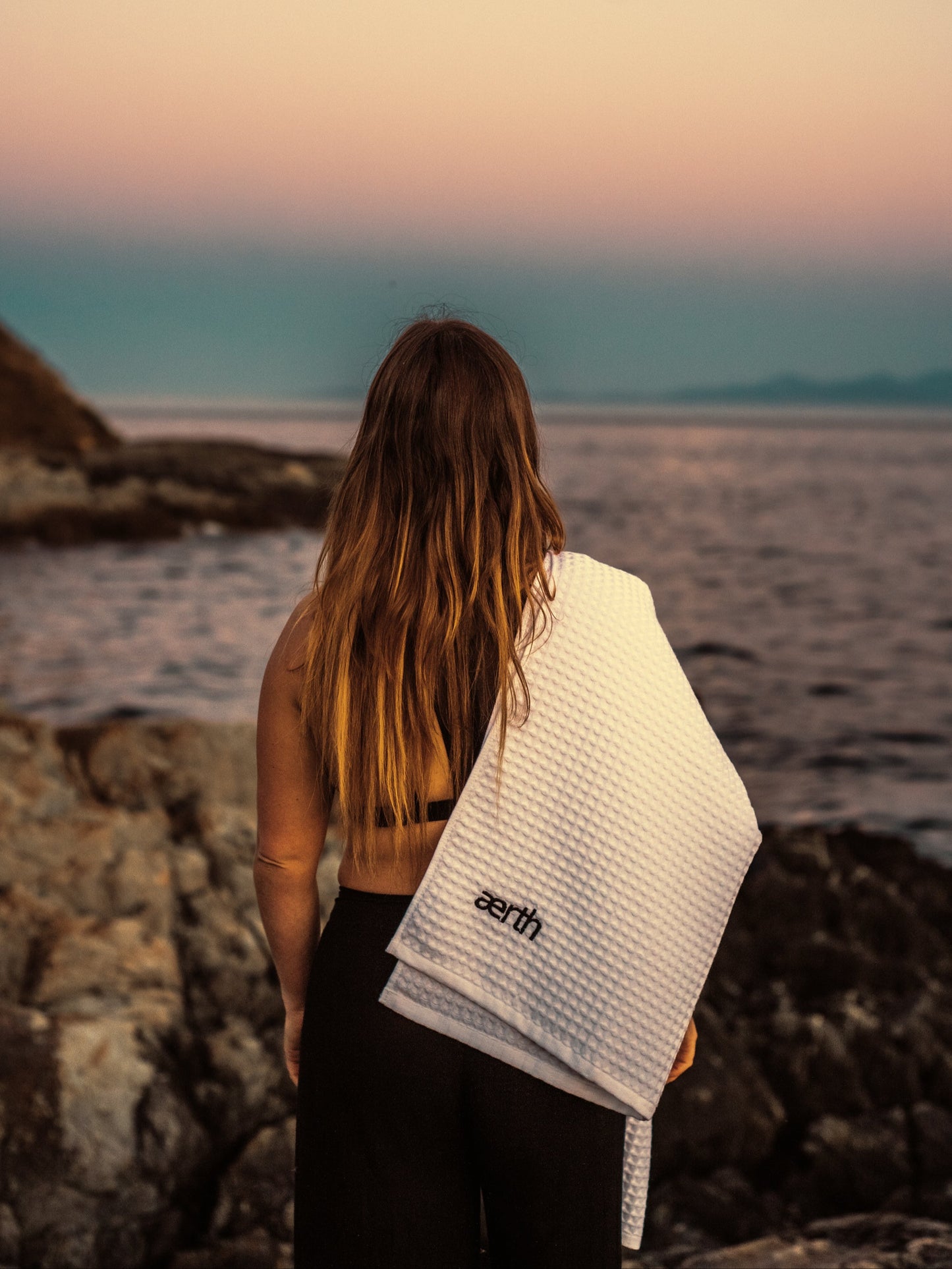 Person standing on a rocky beach with a towel draped over their shoulder, looking out at the ocean.